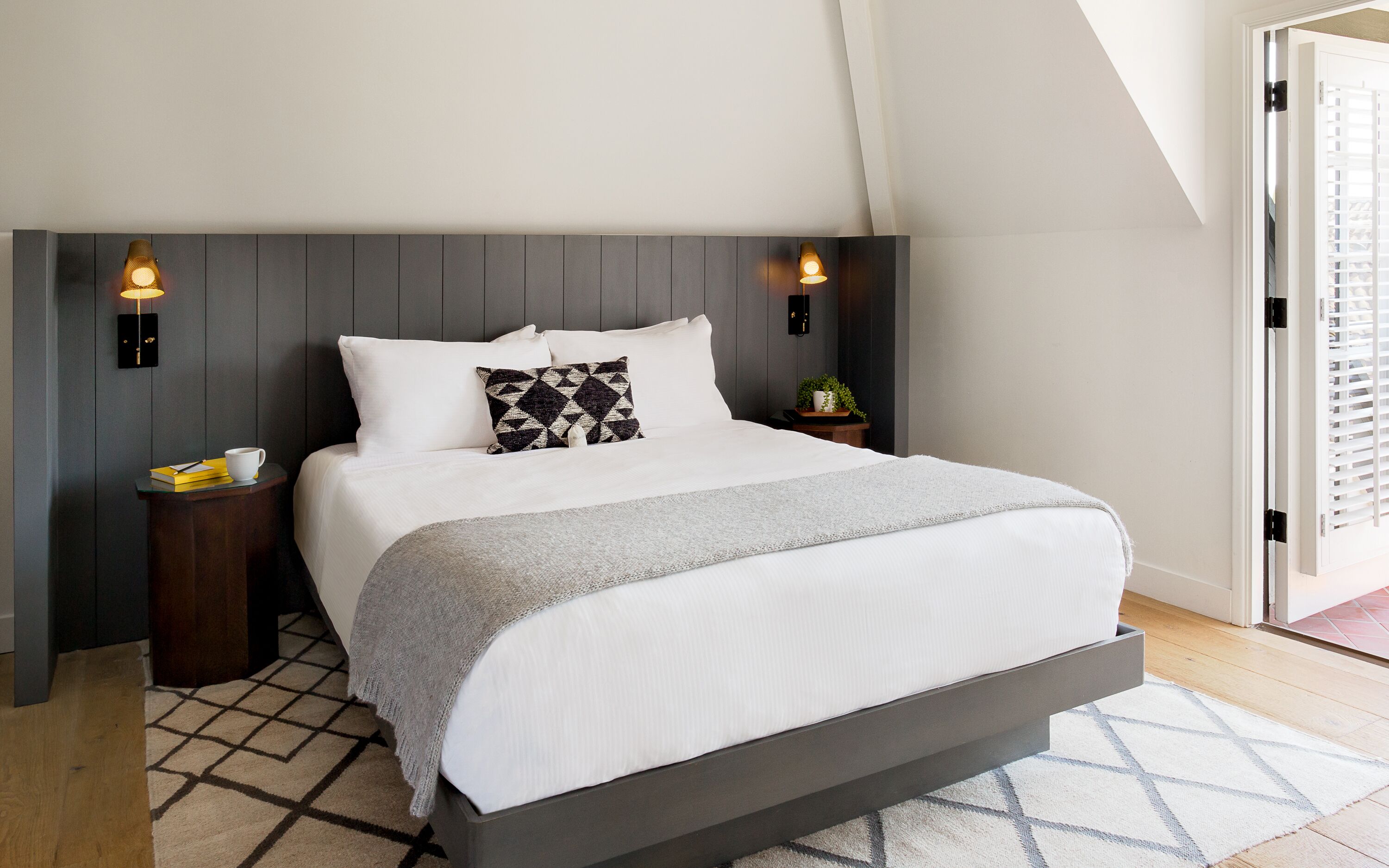 A modern bedroom with a large bed, two bedside tables, a nightstand, a patterned rug, and a view of the outside.