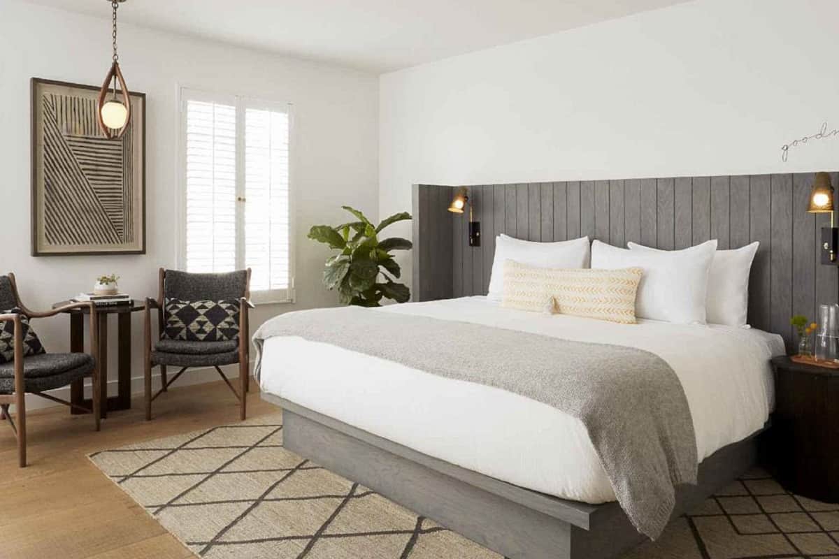 A bedroom with a large bed, a small table, two chairs, a hanging light fixture, and a wall-mounted lamp.