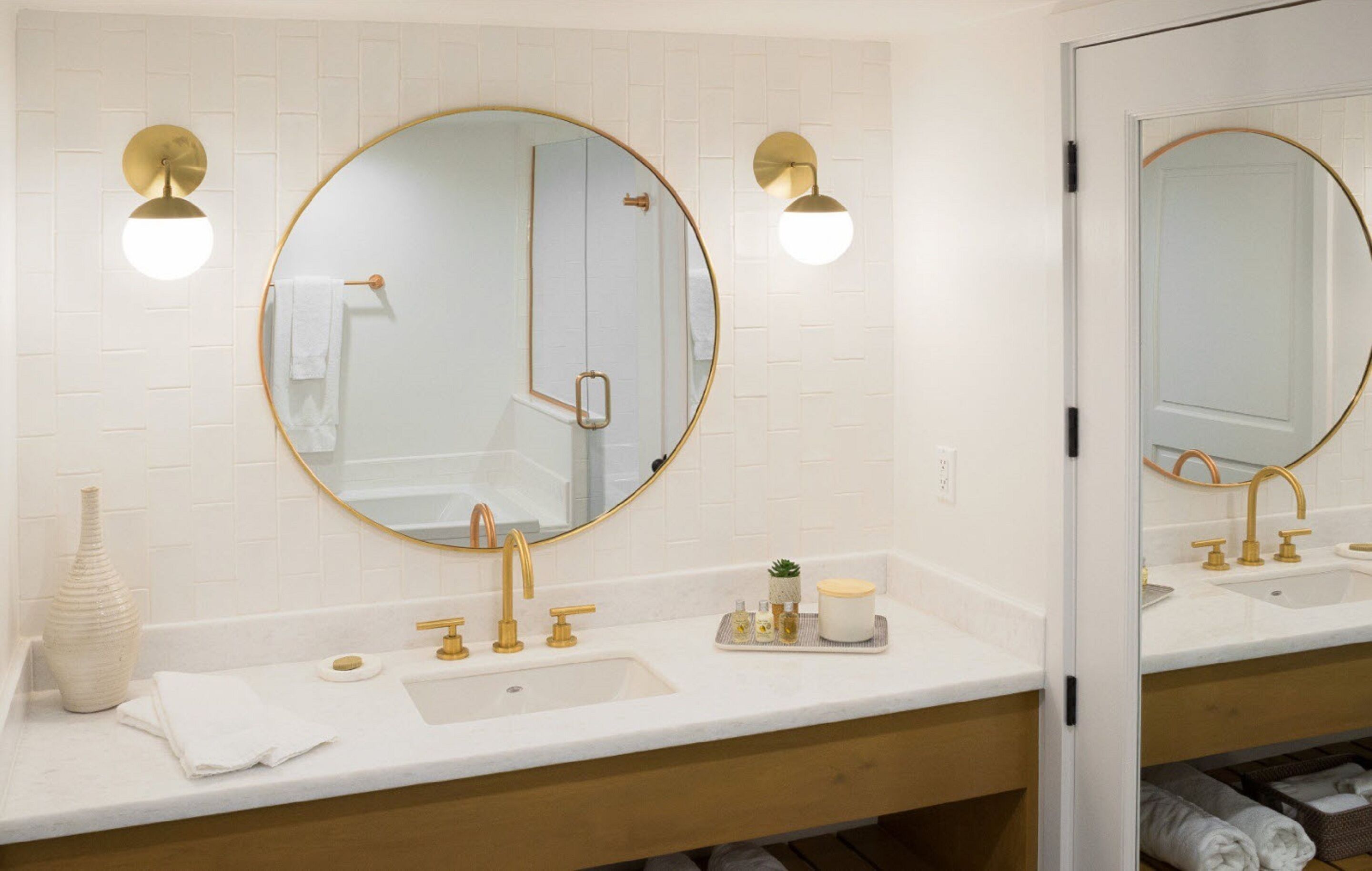 Un baño con dos lavamanos, accesorios dorados, un espejo redondo y una pared de azulejos blancos.