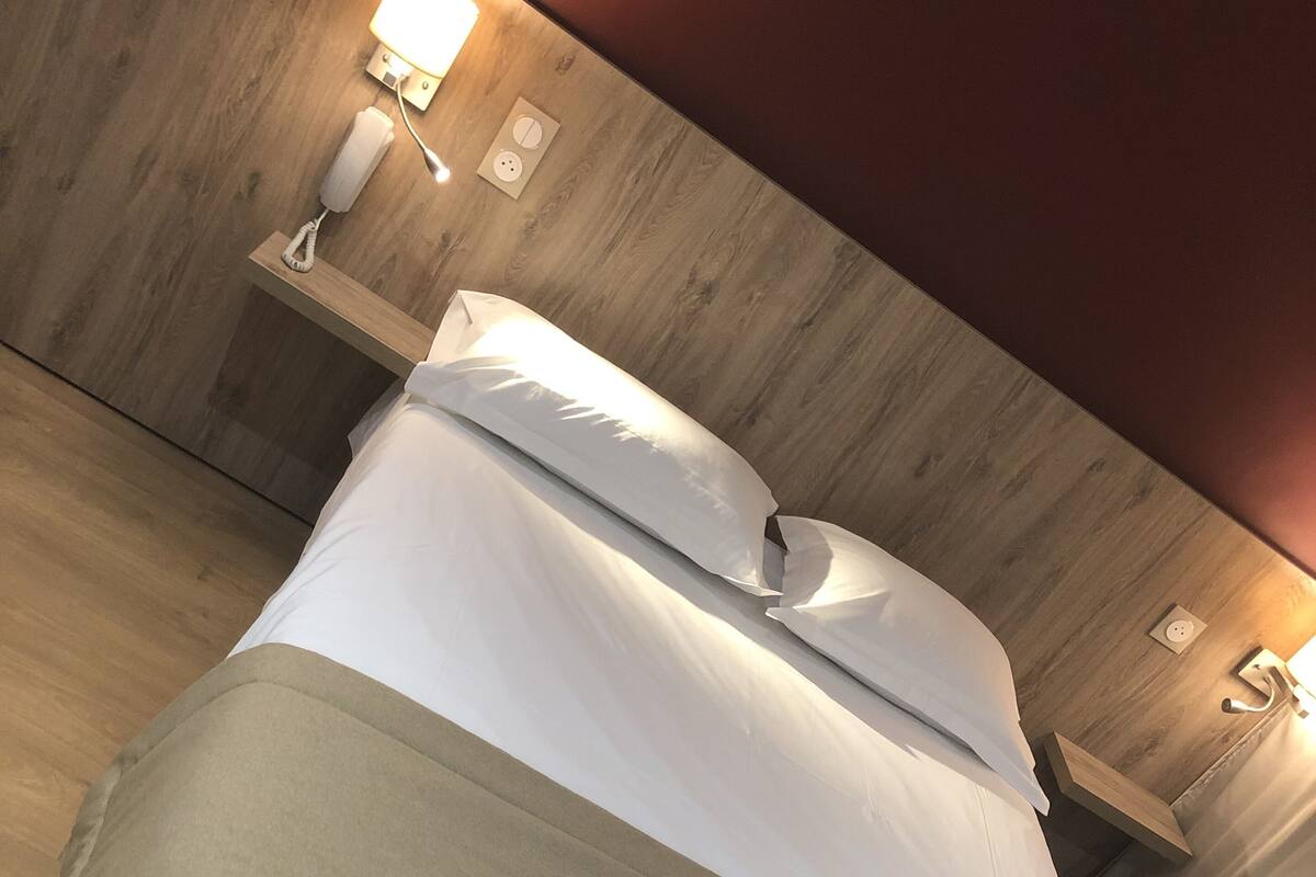 A neatly made bed with a white and beige bedspread, a wooden headboard, and a bedside table with a lamp and a phone.