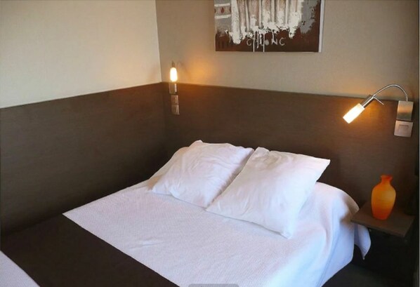 Una cama bien hecha con sábanas blancas y dos lámparas de lectura fijadas a la pared a cada lado.