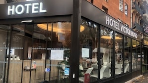 Un hotel con fachada de vidrio moderna, una zona de recepción y asientos exteriores visibles a través de las ventanas.
