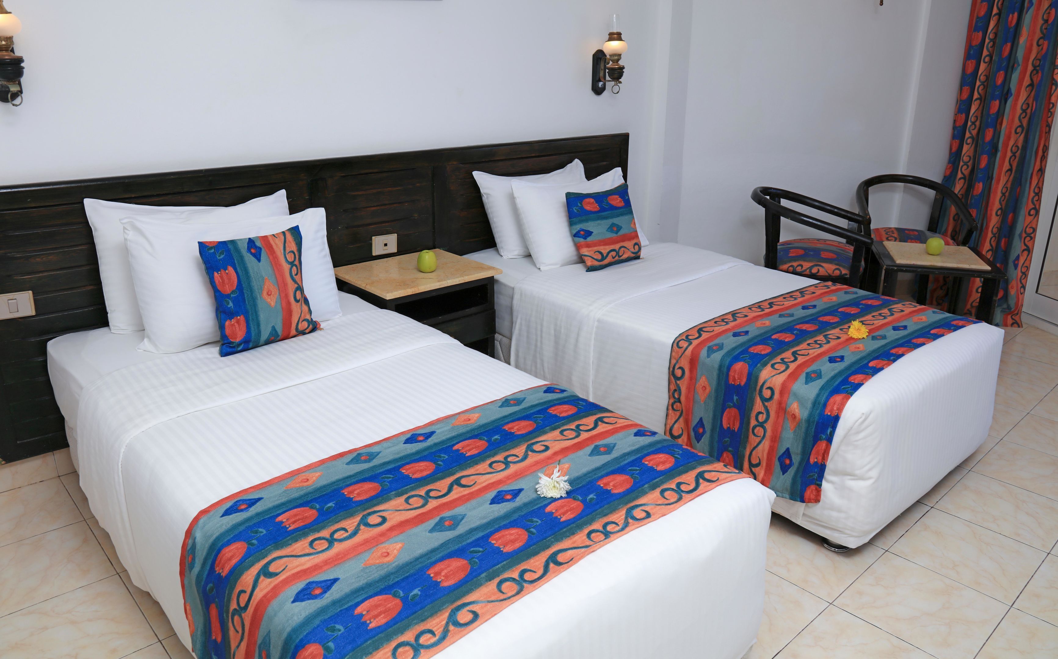 A hotel room with two beds, each with a patterned bedspread, a nightstand, a chair, and a wall-mounted light fixture.