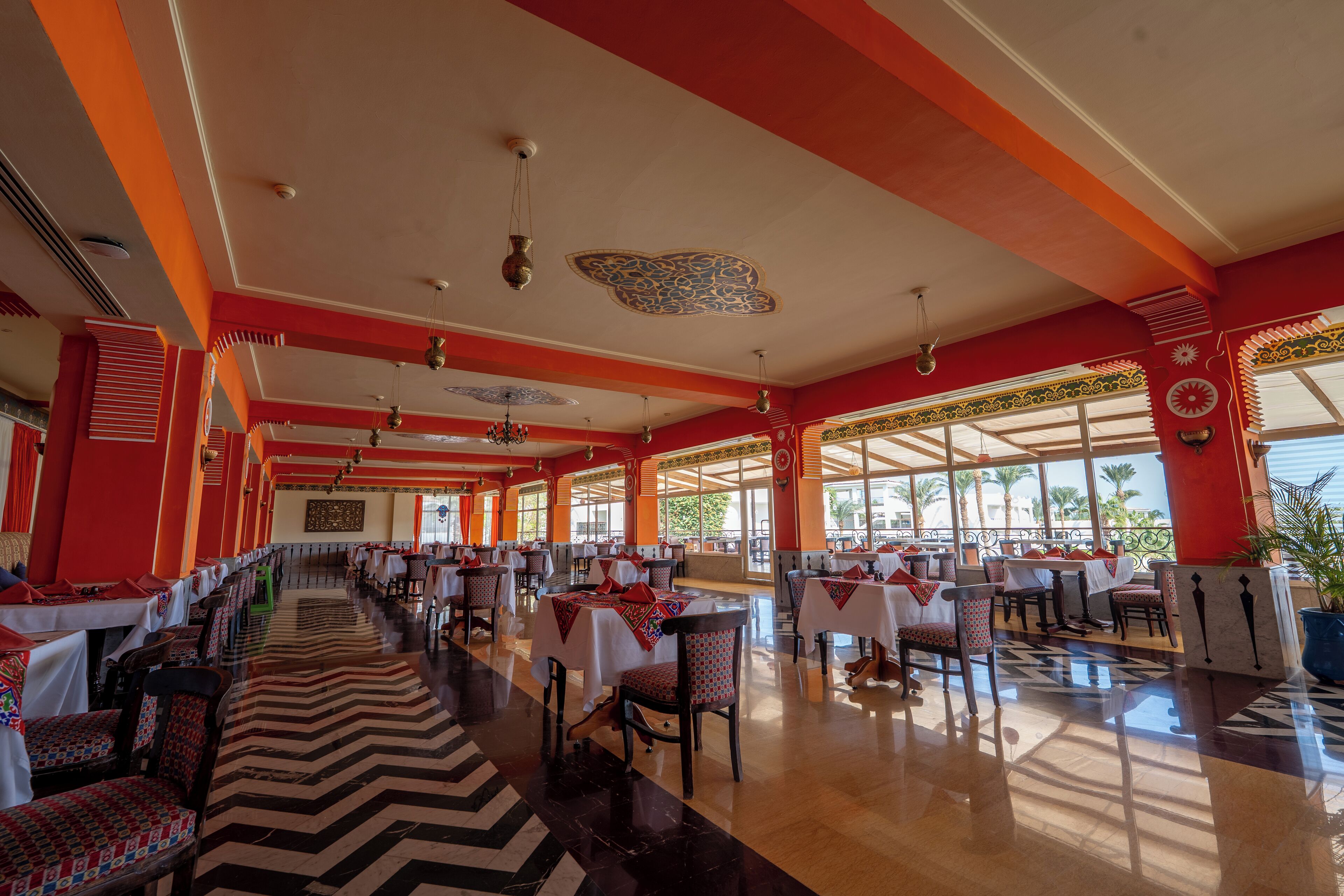 A spacious dining area with multiple tables and chairs, large windows, and a decorative ceiling.