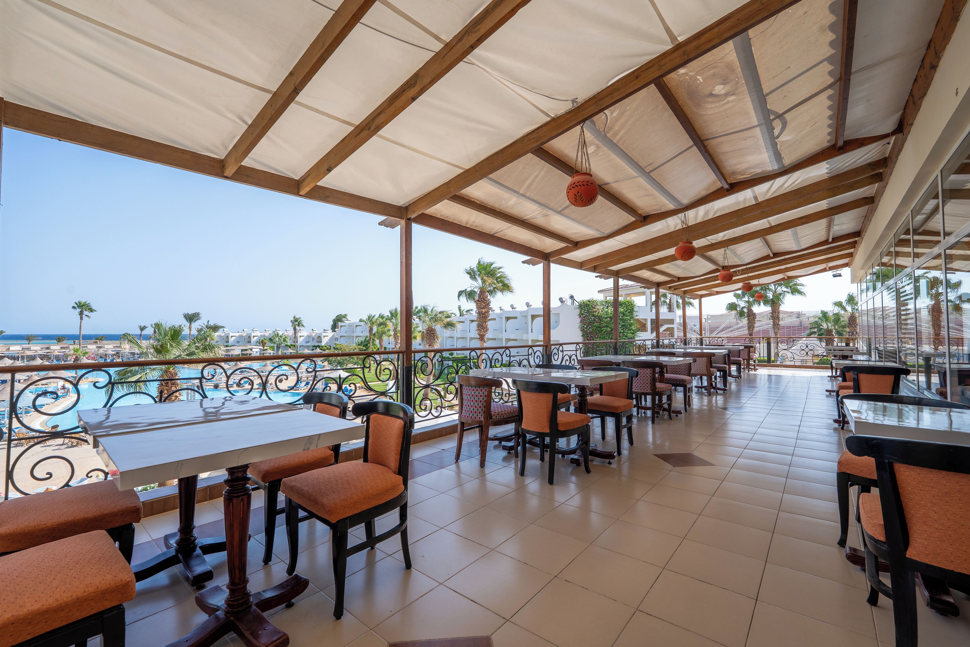 A covered outdoor dining area with tables and chairs, overlooking a pool and palm trees.