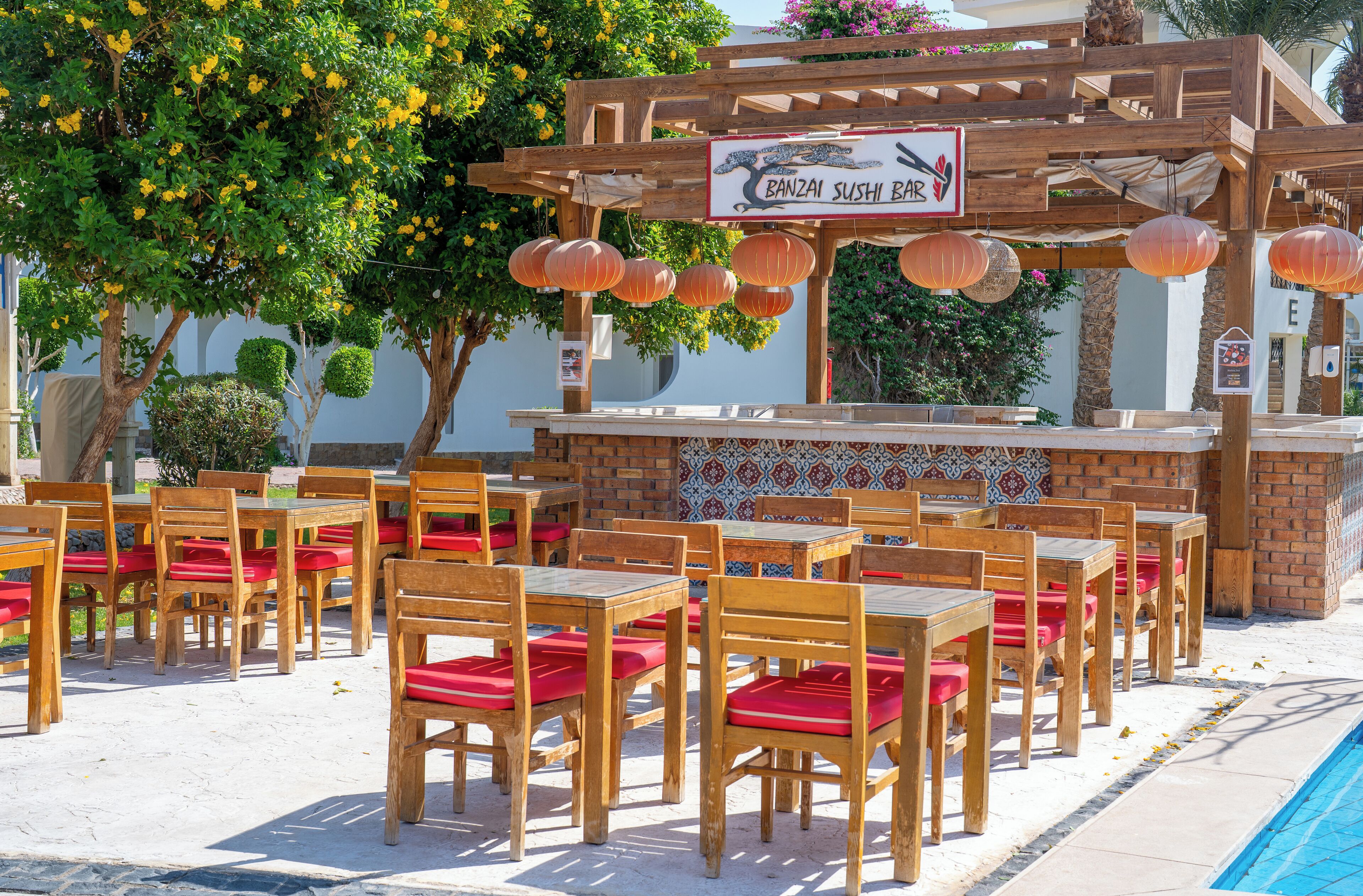 Un restaurant avec une enseigne indiquant « BANZAI SUSHI BAR » et des tables en plein air, en bois, ainsi que des chaises rembourrées de couleur rouge.
