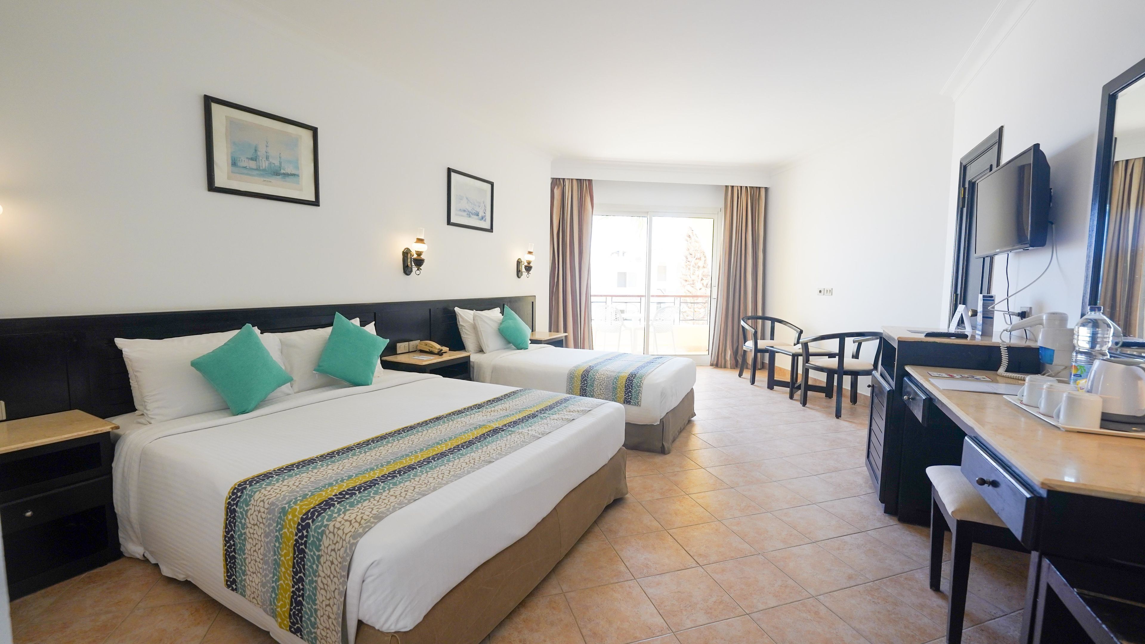 A hotel room with two beds, a desk with a chair, a TV, and a balcony with curtains.