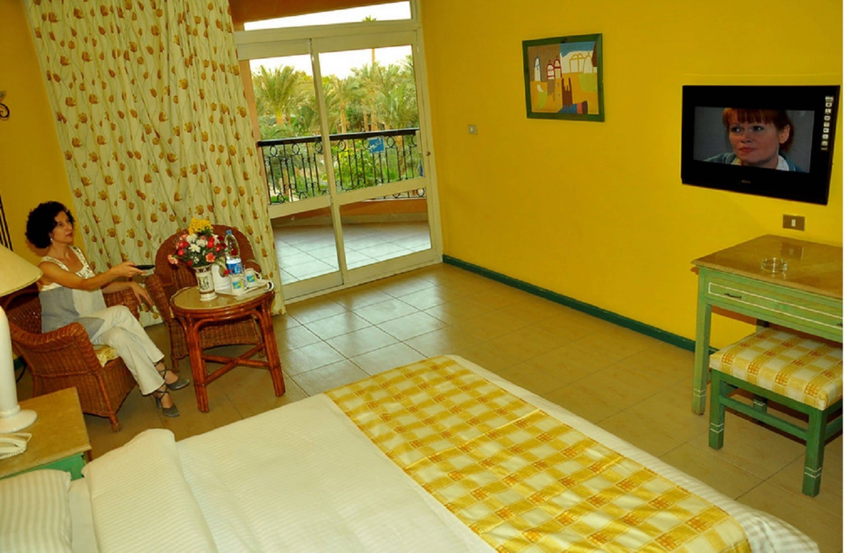 A hotel room with a bed, a television, a balcony with a view of palm trees, and a dining area with a table and chairs.