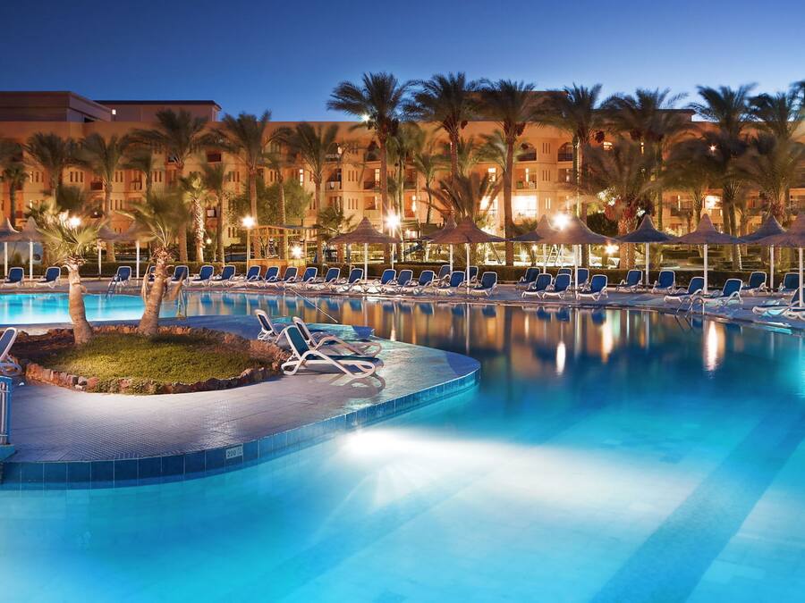 A large outdoor swimming pool with lounge chairs and palm trees, surrounded by hotel buildings.