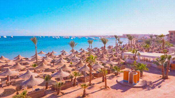 Een strand met veel rieten parasols en palmbomen, een heldere blauwe zee en bootjes in de verte.
