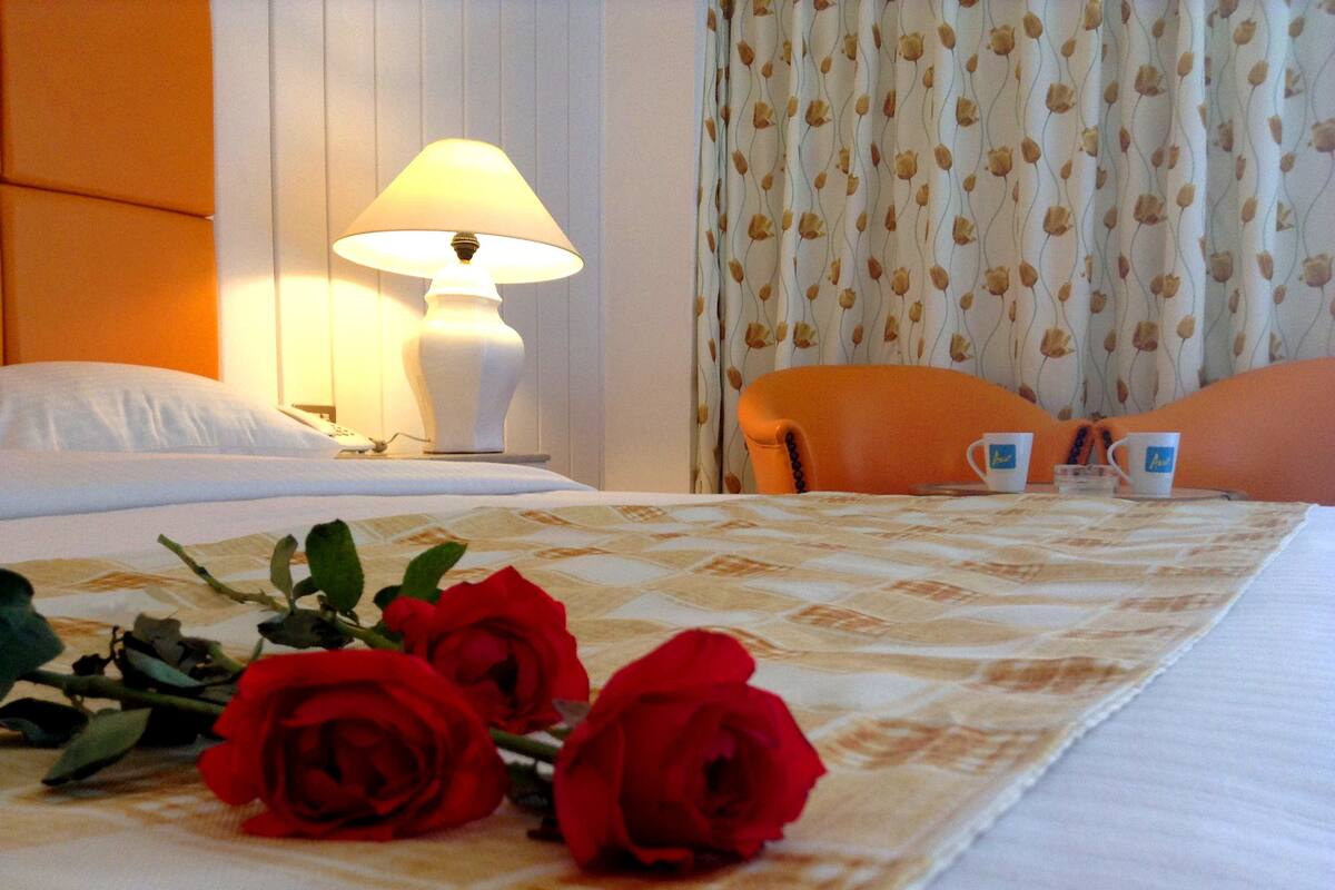 A hotel room with a bed, a bedside lamp, a chair, and a table with two cups.