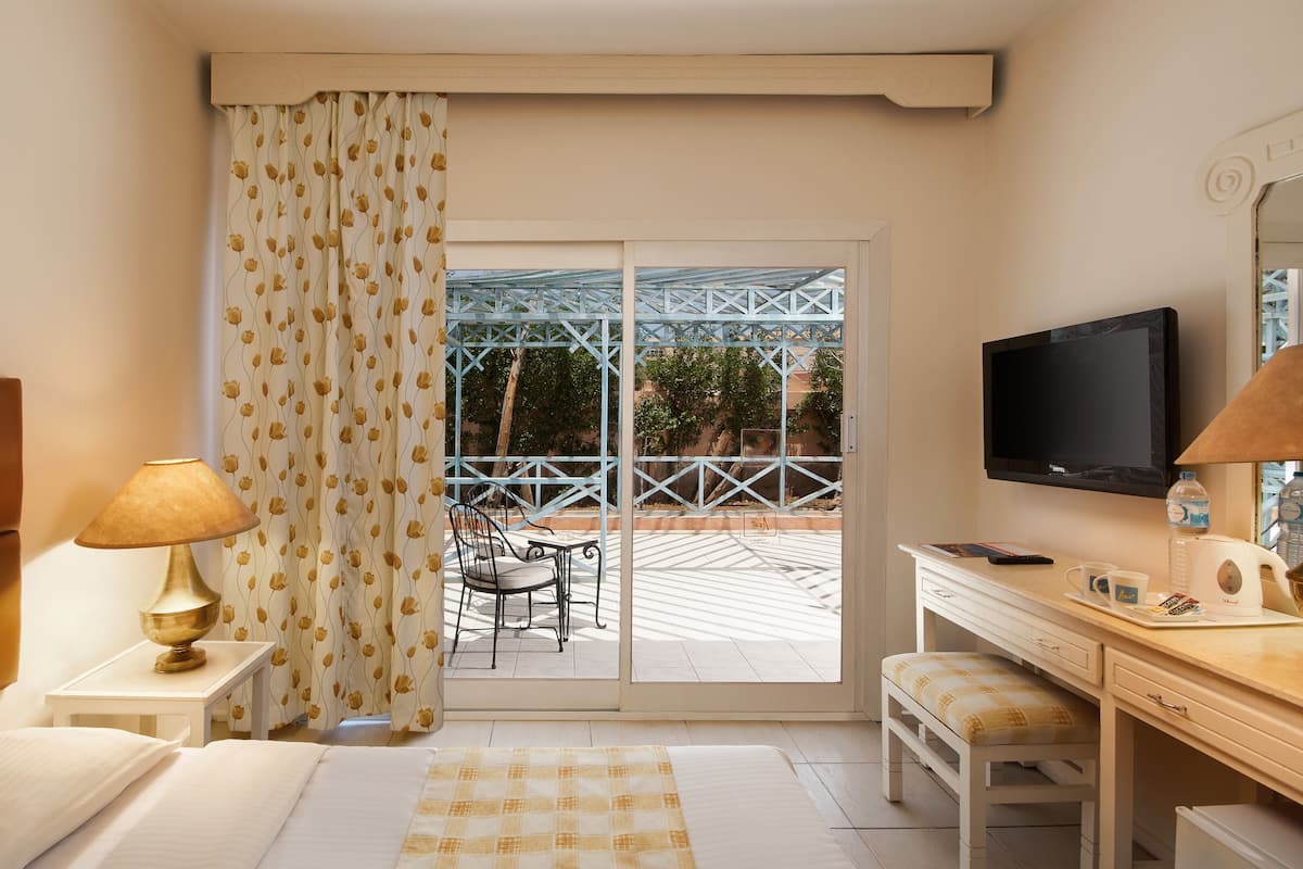 A hotel room with a bed, a desk, a chair, a TV, and a balcony with a view of greenery and a metal structure.
