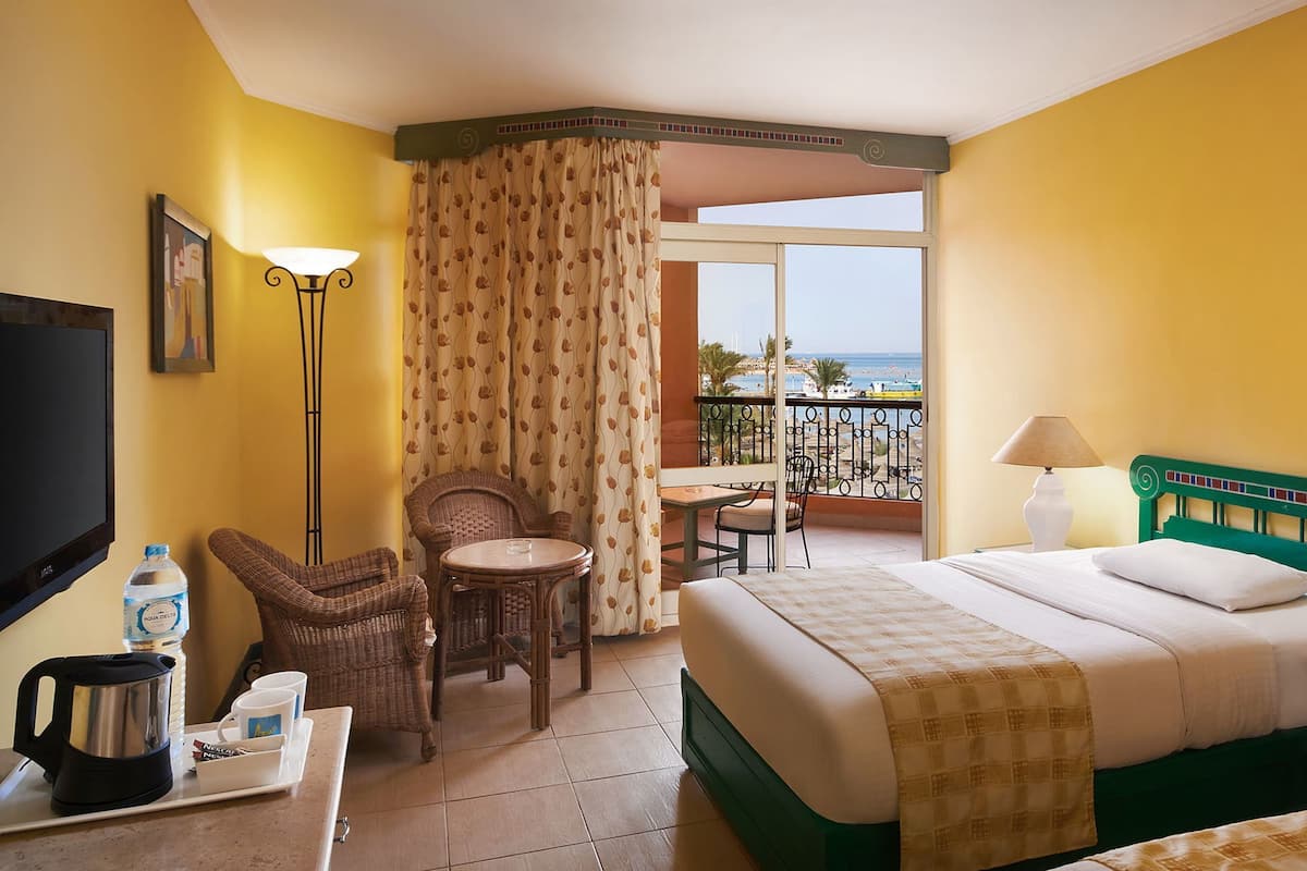 A hotel room with a bed, a desk with a kettle and coffee cups, a chair, a table, a balcony with a view of the sea, and a TV.
