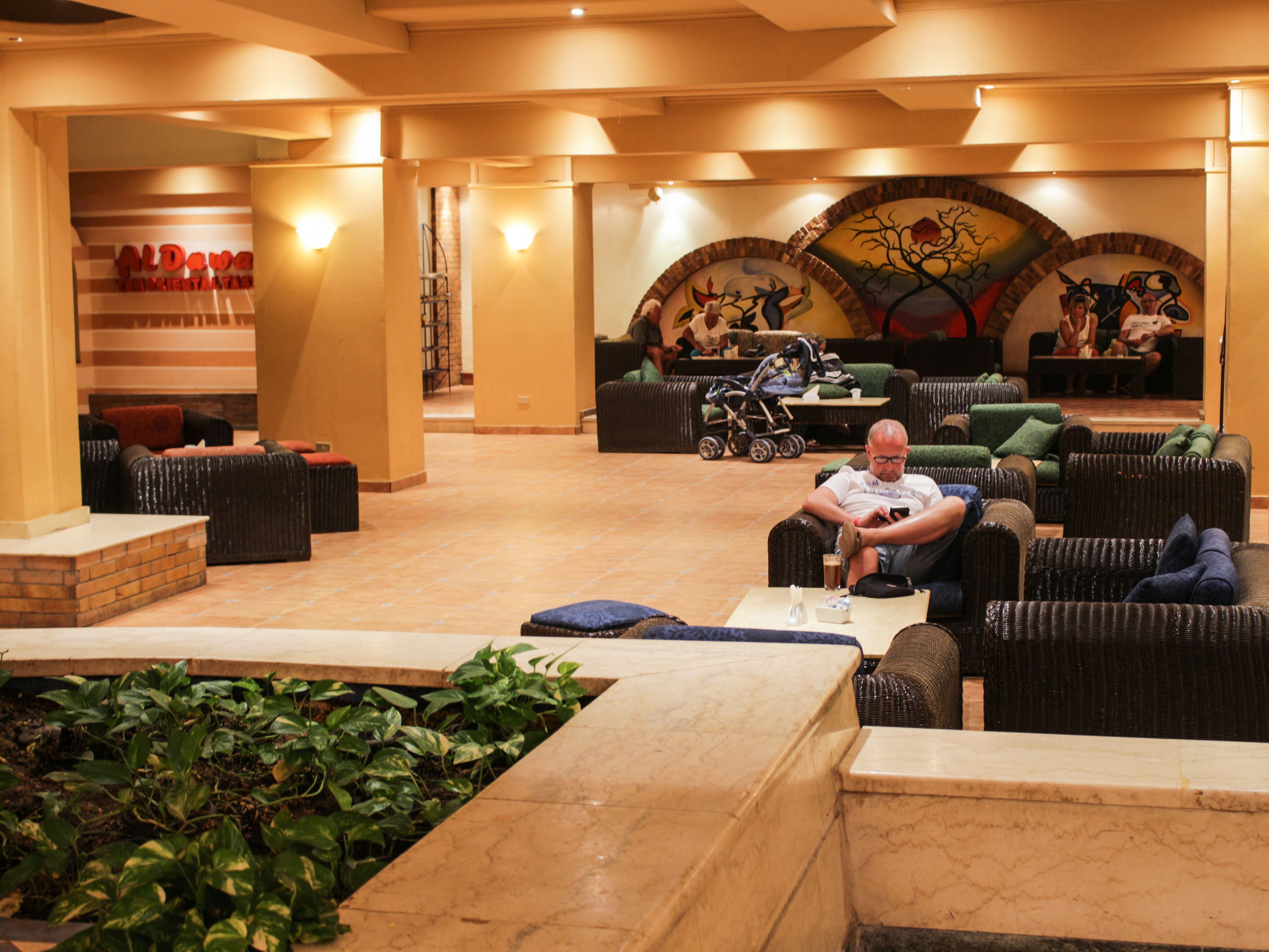 A man seated in a lounge area with wicker furniture, a tiled floor, and a wall with murals.