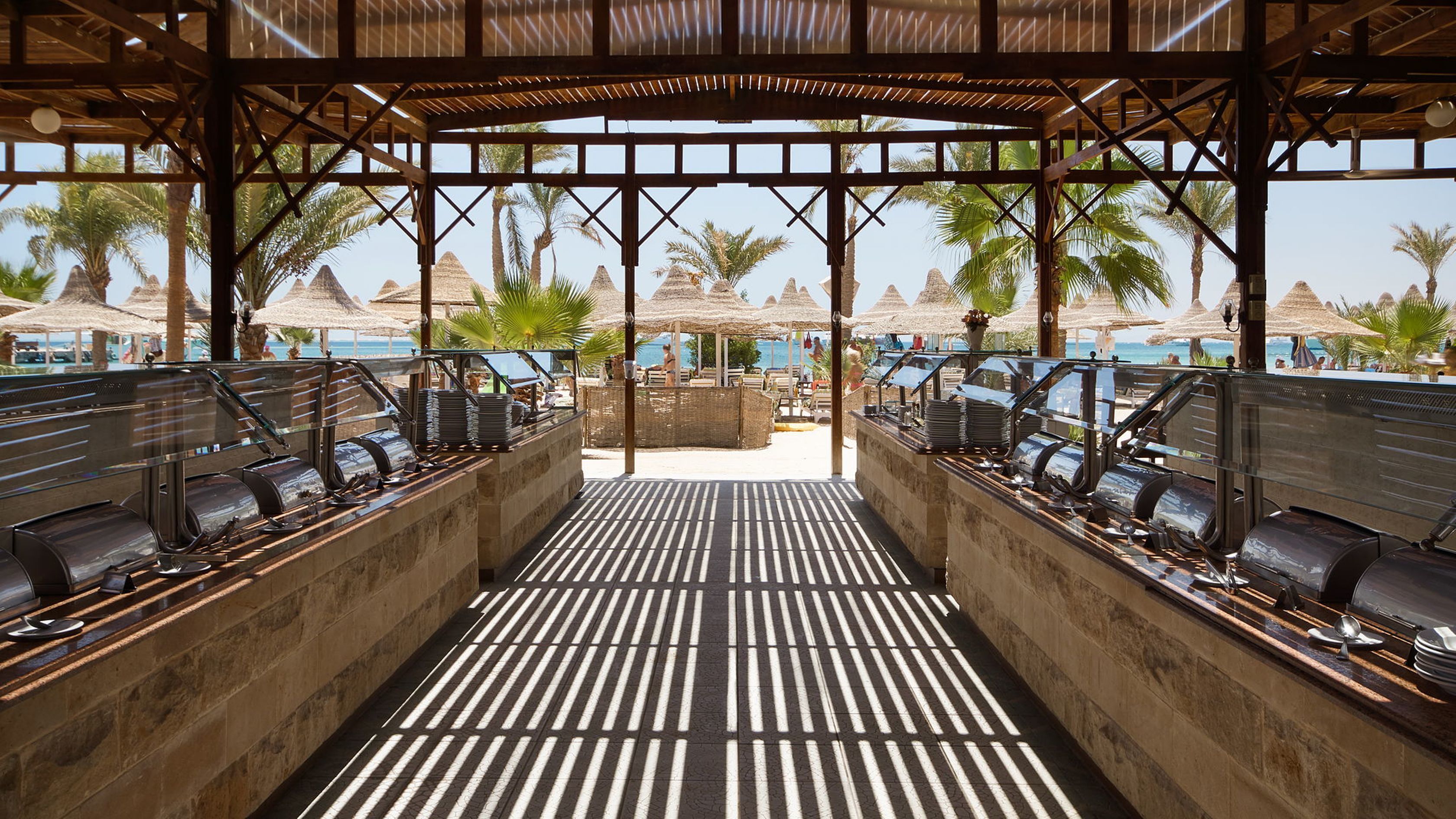 A covered outdoor dining area with a central walkway, surrounded by palm trees and beach umbrellas.