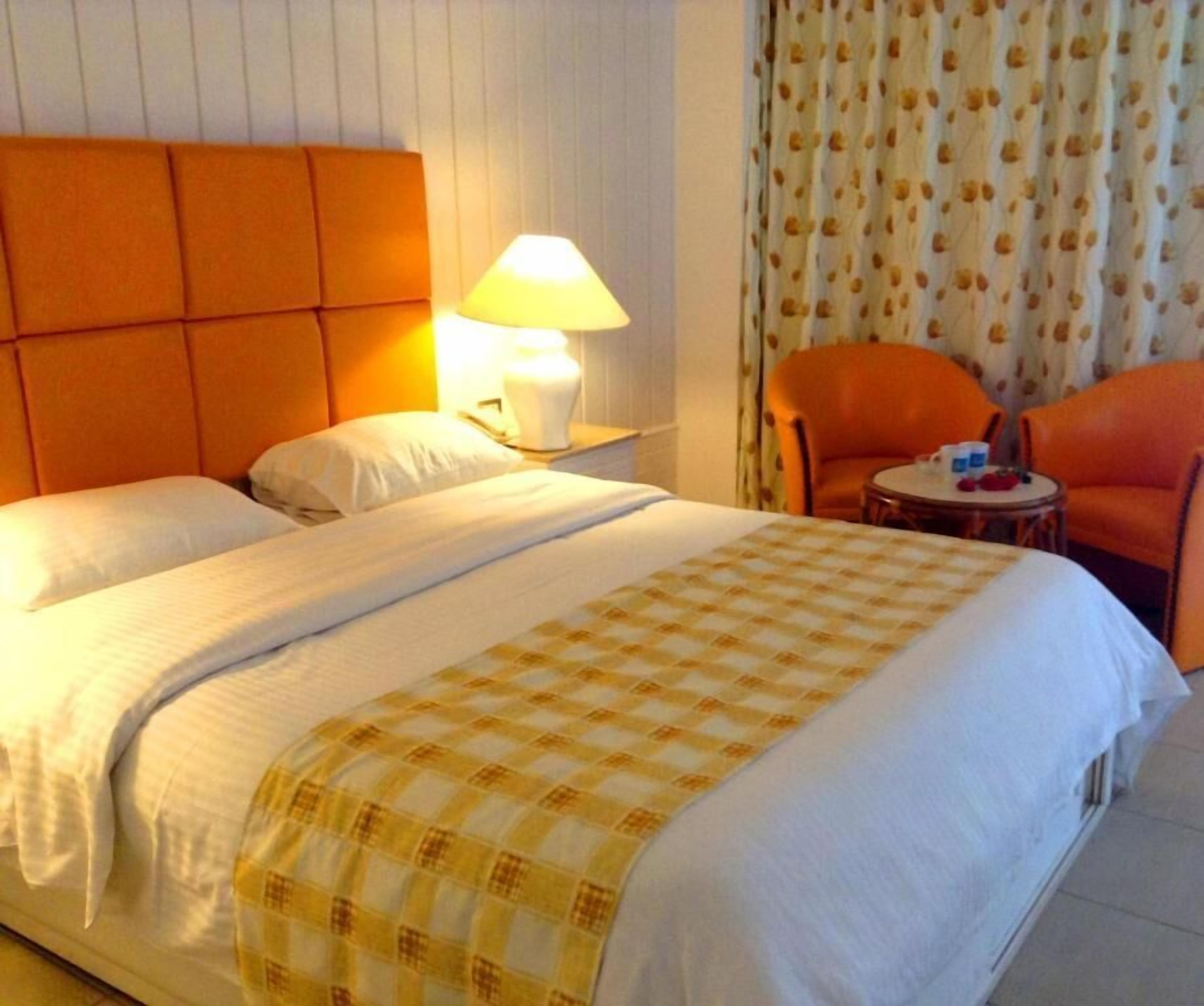 A hotel room with a bed, a nightstand with a lamp, two orange chairs, and a small table with items on it.