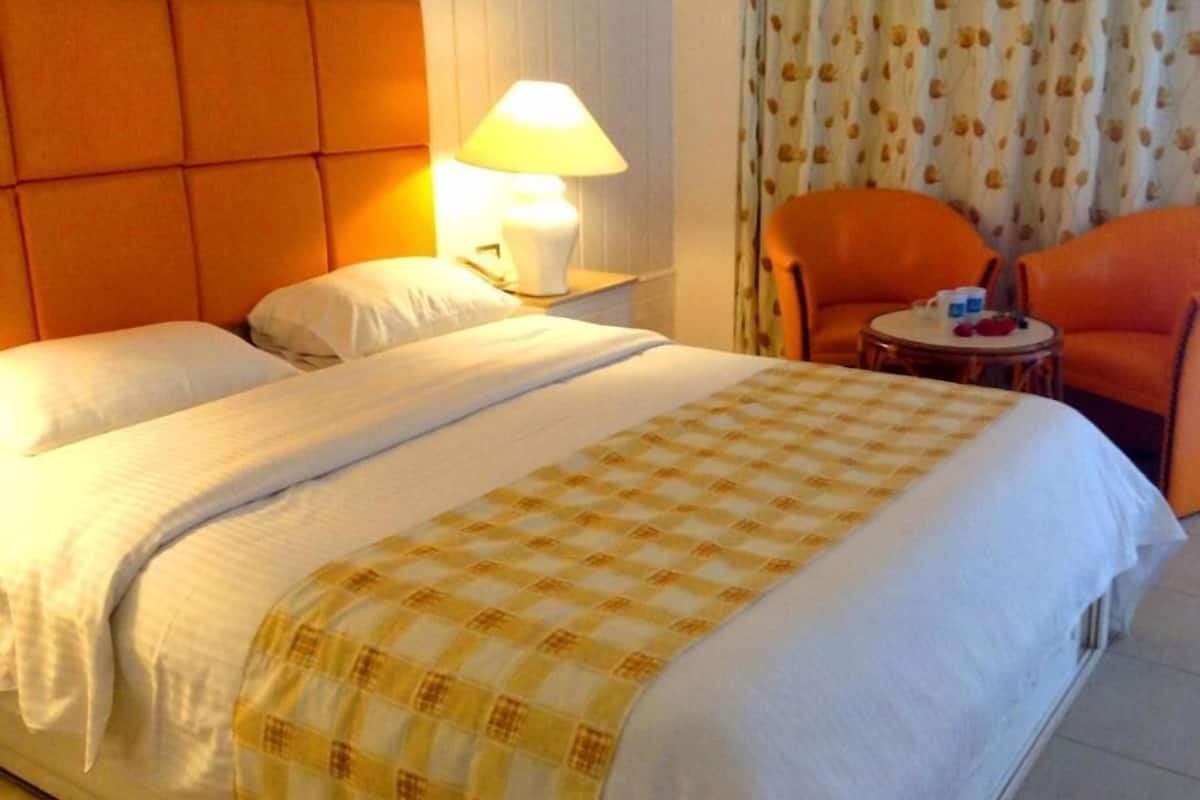 A hotel room with a bed, a nightstand with a lamp, two orange chairs, and a small table with items on it.