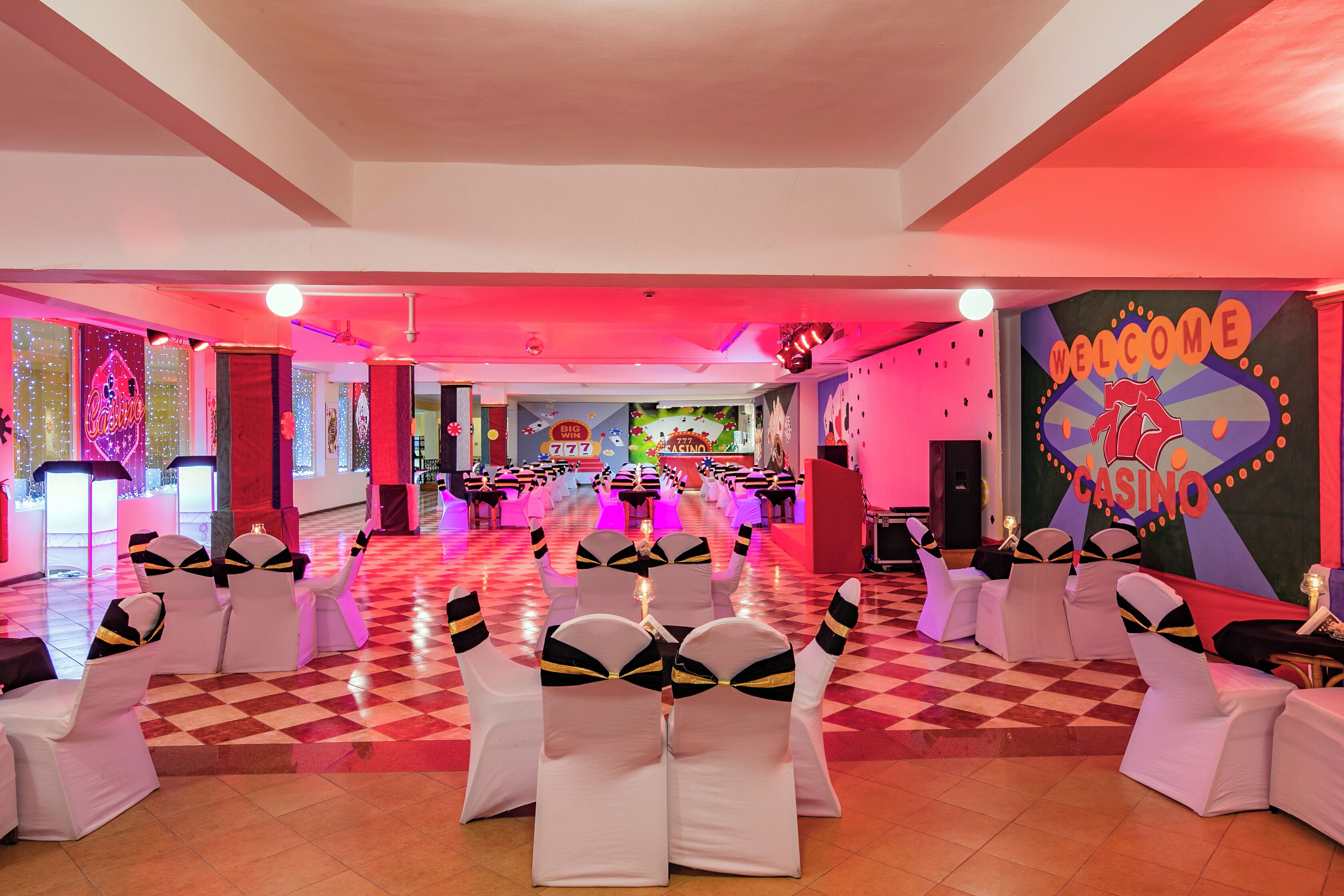 Ein geräumiger Saal mit Reihen weißer Stühle mit schwarzen und goldenen Bändern, einem Schachbrettfußboden und einem Schild an der Wand mit der Aufschrift 'Welcome to the Casino'.