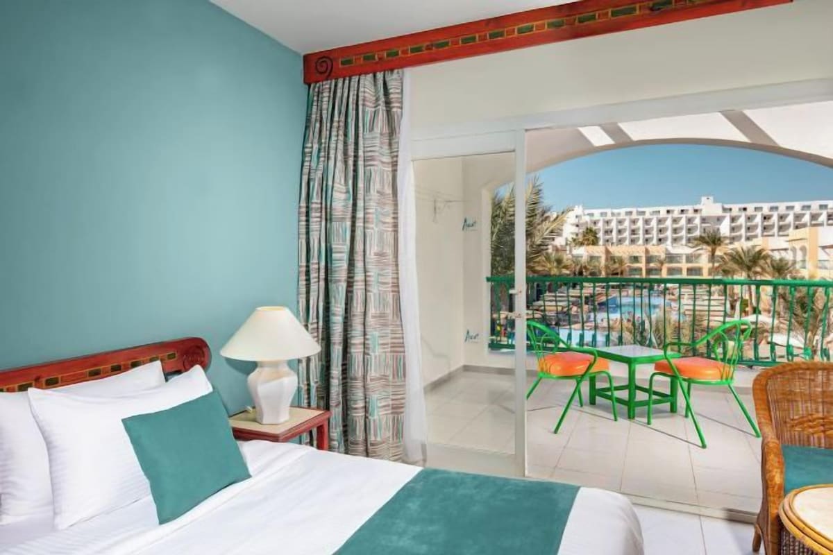 Habitación de hotel con cama, mesita de noche, lámpara y balcón con vistas a una piscina y un edificio.