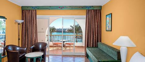 Een hotelkamer met een bed, een bank, een tafel en een stoel. Er is een balkon met uitzicht op een zwembad en palmbomen.
