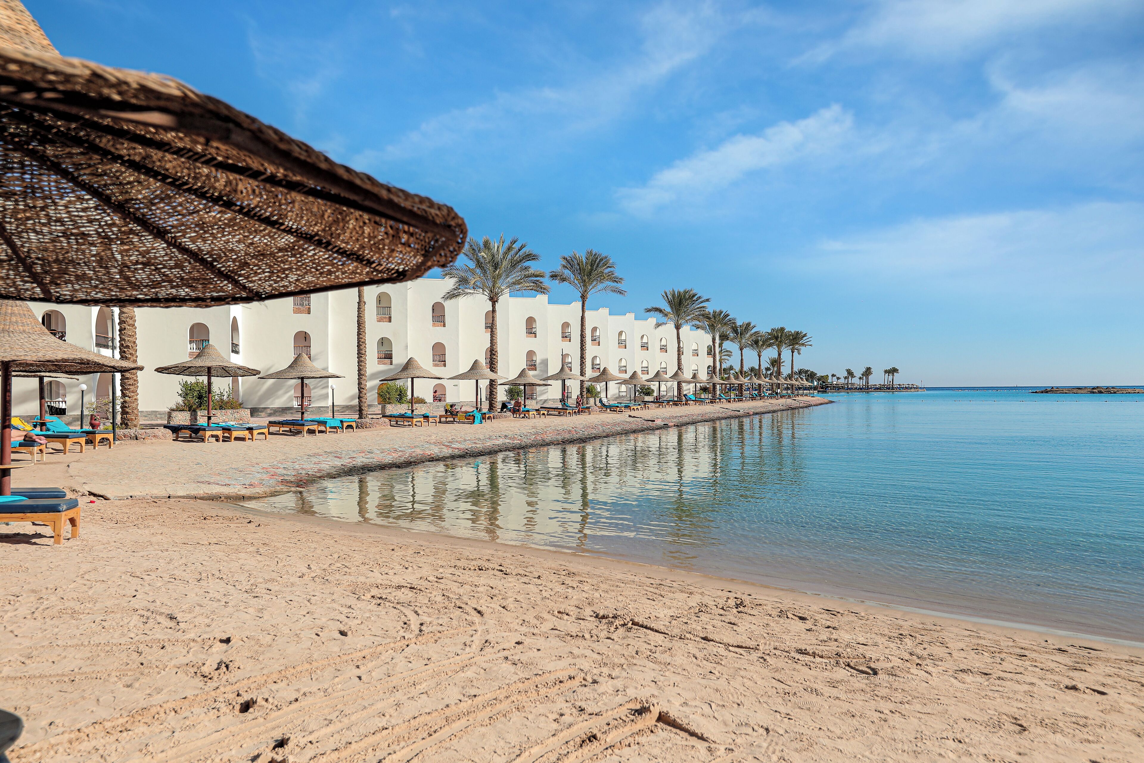 A beach with sun loungers and umbrellas, a water body, and a row of white buildings with arches.