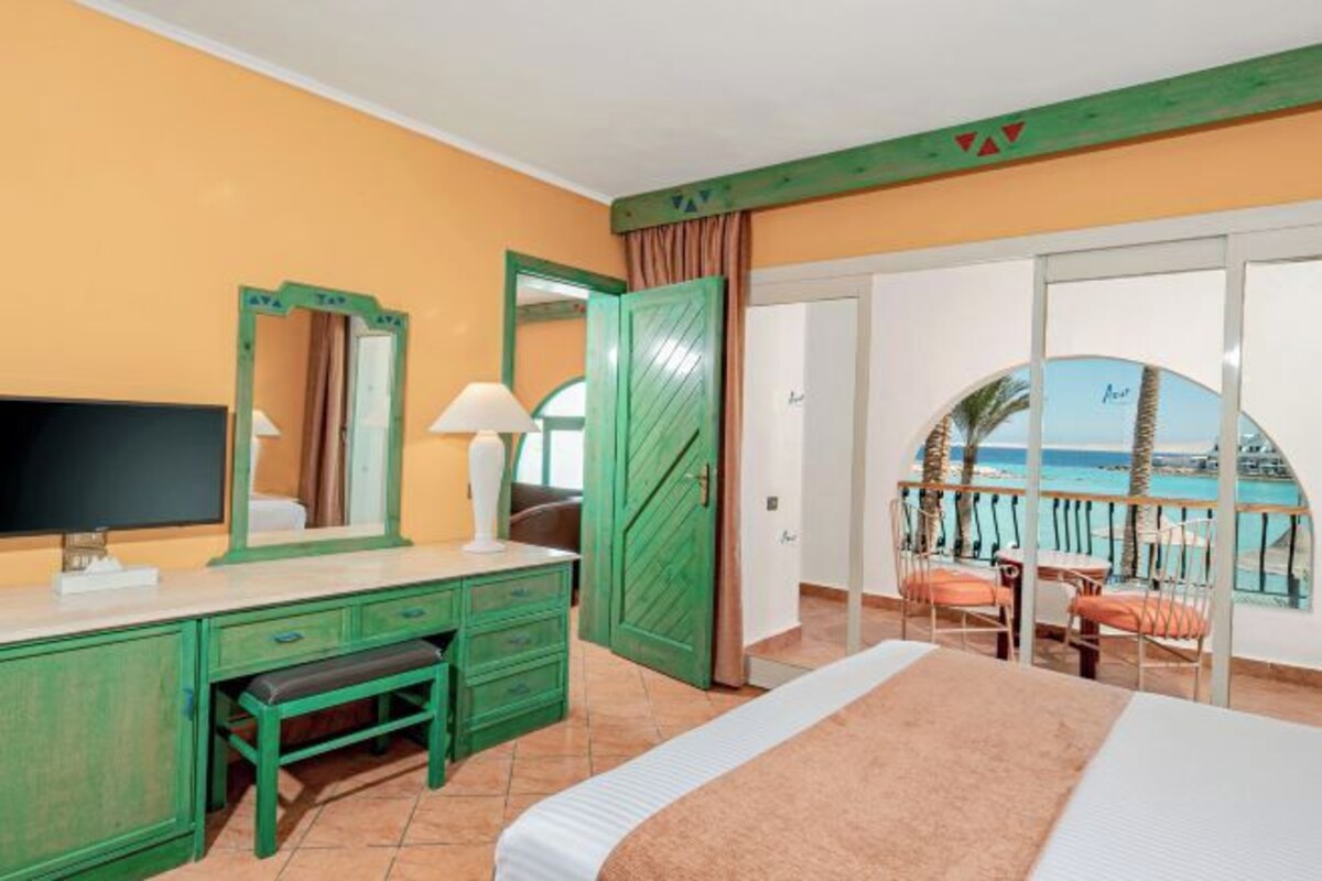 A hotel room with a bed, a desk with a mirror, a television, and a view of the beach.