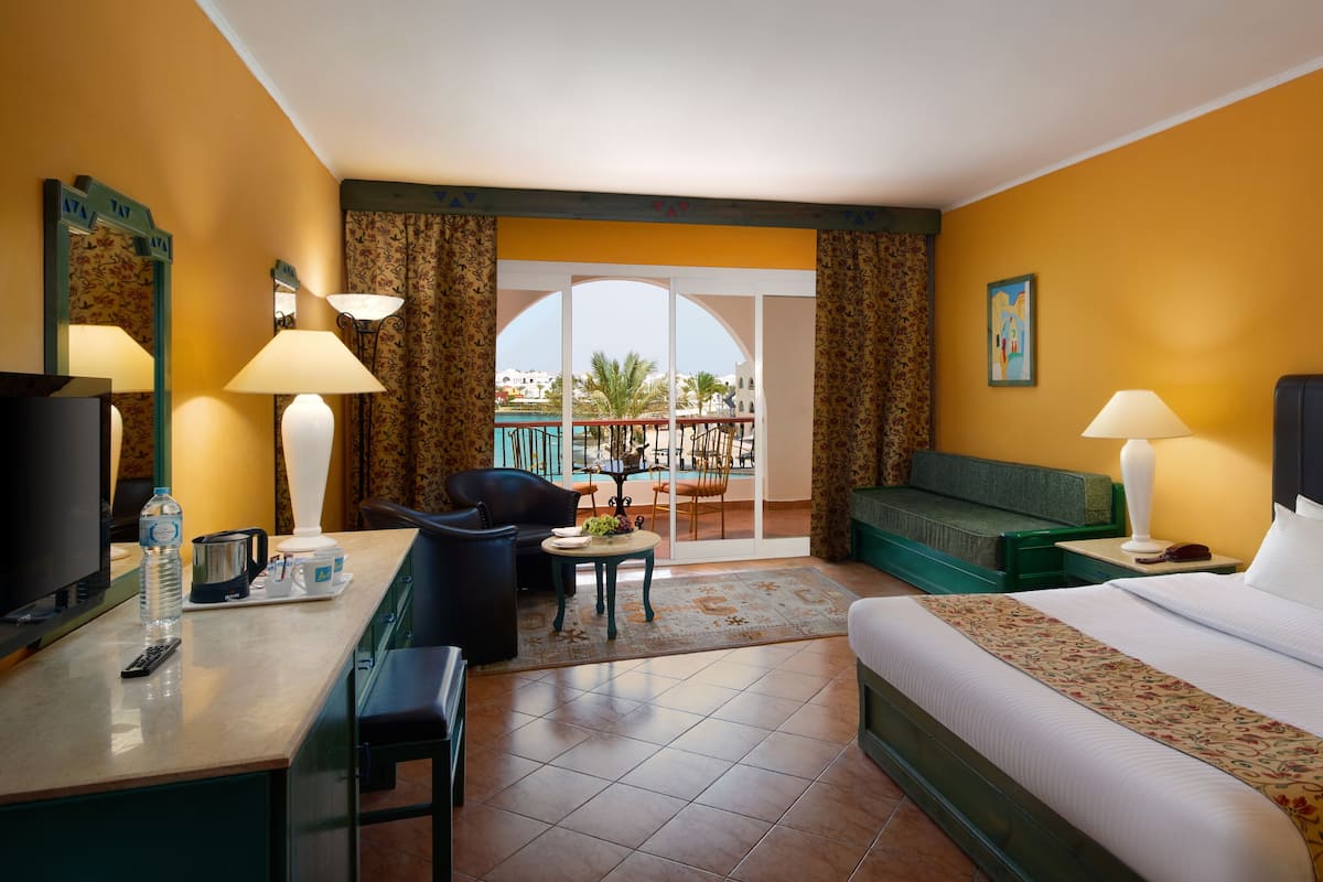 A hotel room with a bed, a desk with a TV, a chair, a sofa, a table with a lamp, and a view of a pool and palm trees.