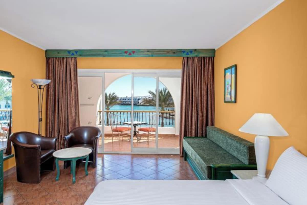 A hotel room with a balcony, a bed, a sofa, a chair, a small table, and a lamp.