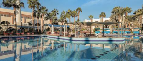 A swimming pool with lounge chairs and umbrellas, surrounded by palm trees and a building with arches.