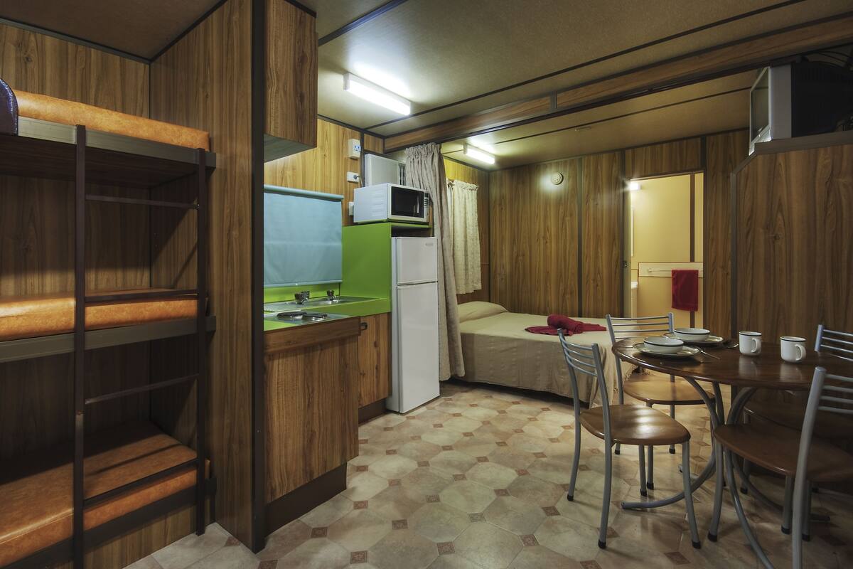 A compact kitchen with a sink, microwave, and refrigerator. There is a dining table with chairs and a bed in the adjacent room.