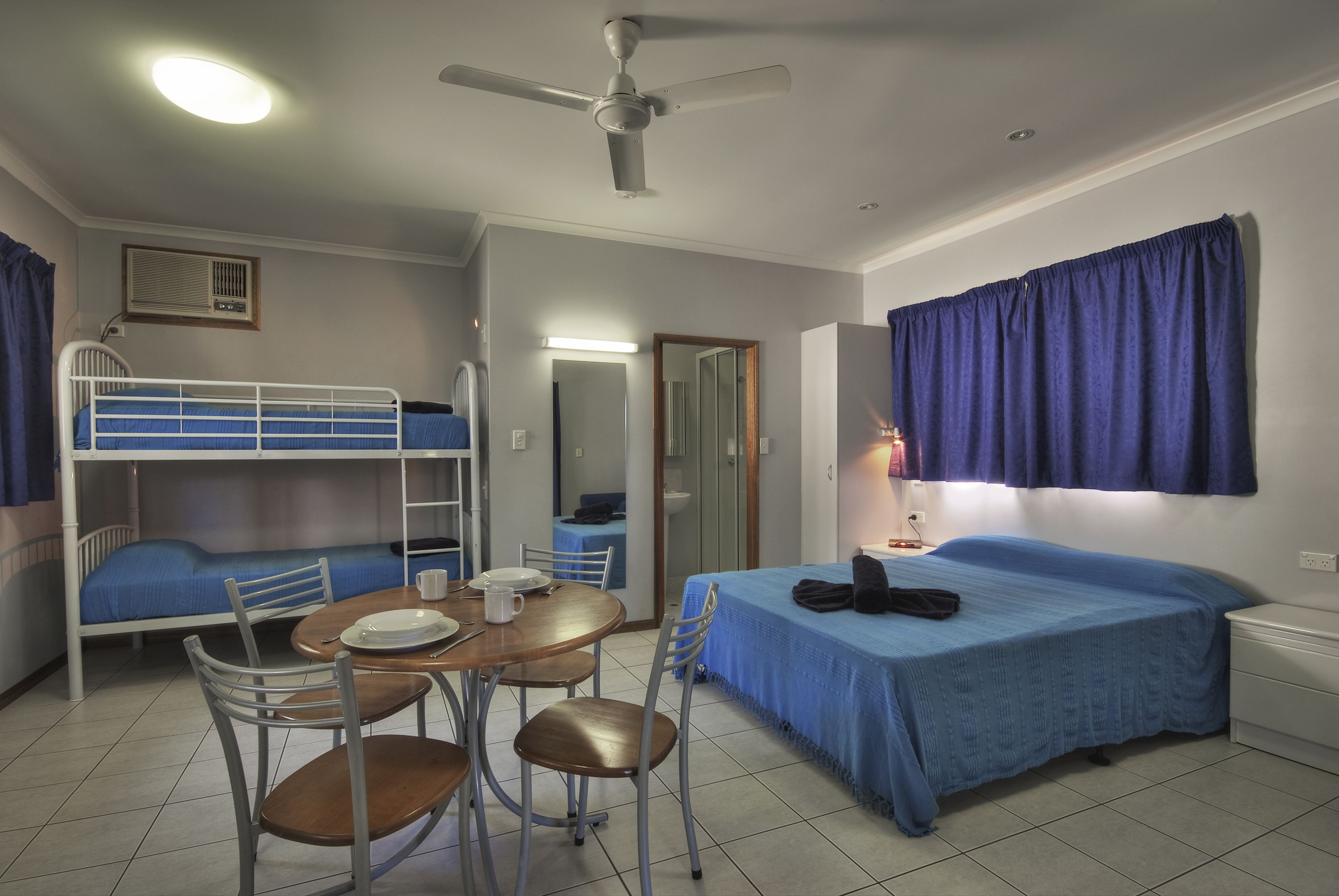 A dormitory room with bunk beds, a single bed, a table with chairs, a ceiling fan, and a window with curtains.
