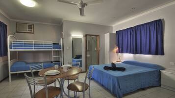 A dormitory room with bunk beds, a single bed, a table with chairs, a ceiling fan, and a window with curtains.