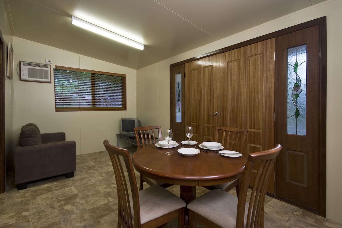A dining area with a round wooden table set for four, surrounded by wooden chairs with cushioned seats. A sofa and a television are visible in the background.