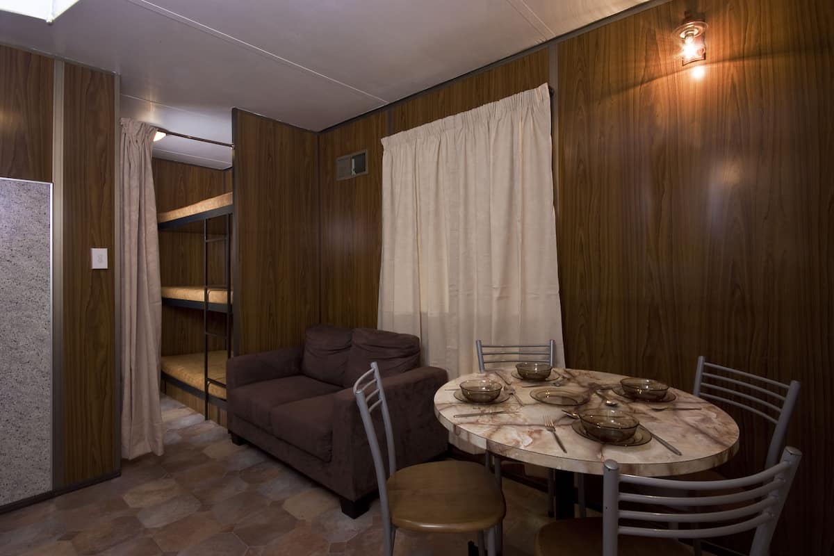 A compact room with a bunk bed, a sofa, a dining table with chairs, and a wooden wall.