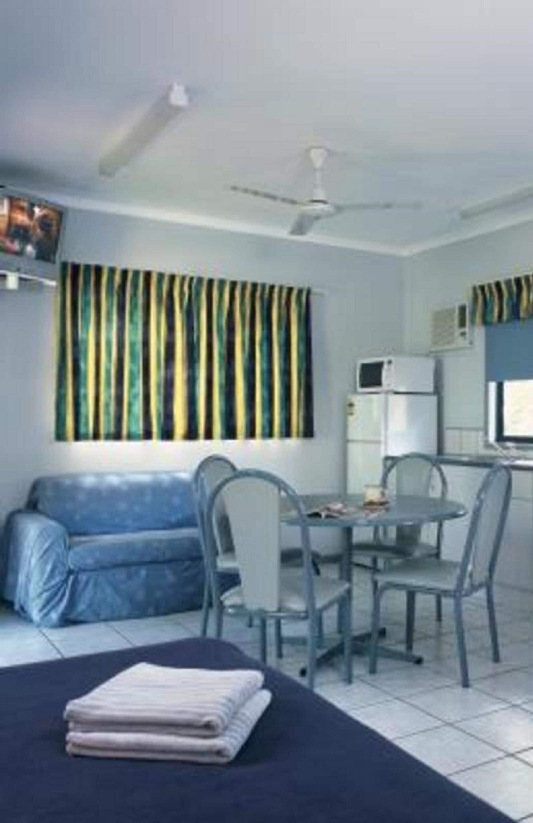 A compact hotel room with a bed, a small dining table with chairs, a sofa, a ceiling fan, a microwave, and a window with striped curtains.