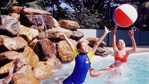 Two people playing with a beach ball in a pool surrounded by rocks and palm trees. - Cairns Sunland Leisure Park (Manoora)