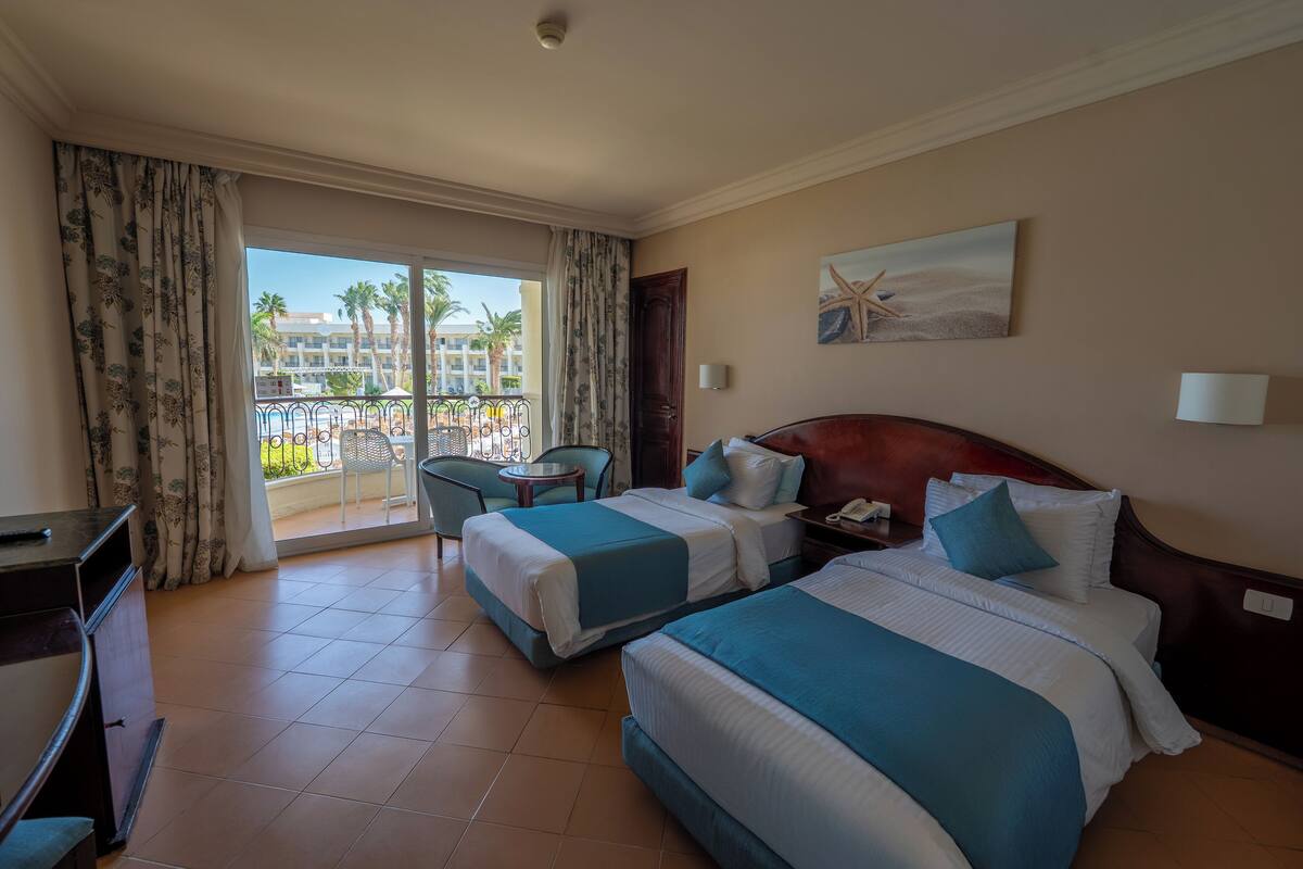 Camera d'albergo con due letti, una scrivania, una sedia e un balcone con vista su palme e edifici.