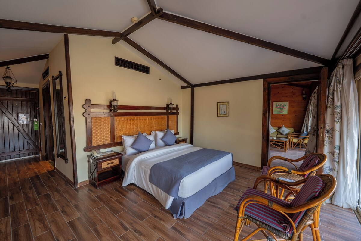 Una camera da letto con un letto grande, pavimento in legno e soffitto in legno. Sono presenti due poltrone in vimini e un tavolino con una lampada.