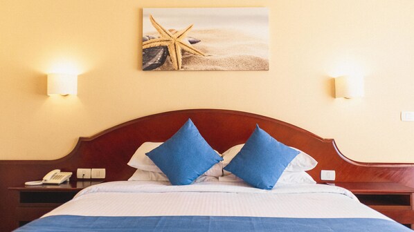 Habitación de hotel con cabecera de madera, cama con ropa de cama azul y blanca, dos almohadas azules, mesita de noche con teléfono y un cuadro colgado en la pared de una estrella de mar y una concha.