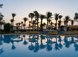 Un área junto a la piscina con sillones de descanso y sombrillas, rodeada de palmeras.