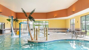 An indoor swimming pool with a water slide, palm trees, and a seating area with tables and chairs.