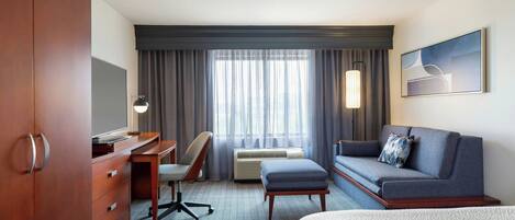 A hotel room with a wooden wardrobe, a desk with a chair, a sofa, a large window with curtains, and a painting on the wall.