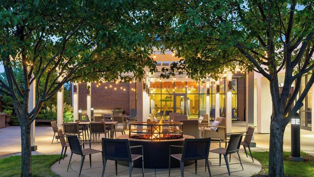 An outdoor patio area with a circular fire pit surrounded by chairs, a brick building in the background, and string lights.