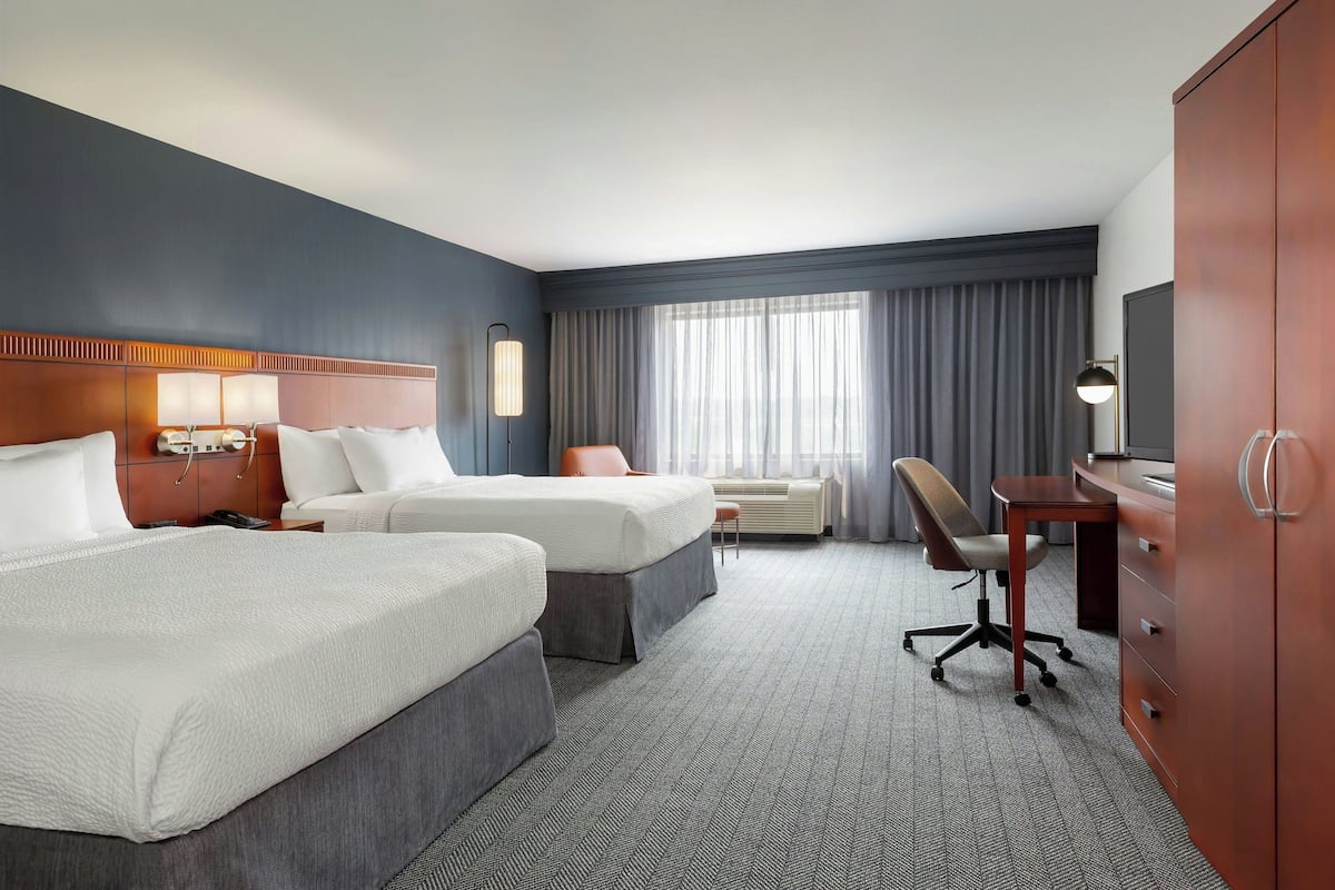 Ein Hotelzimmer mit zwei Betten, einem Schreibtisch, einem Stuhl, einem Kleiderschrank und einem großen Fenster mit Vorhängen.