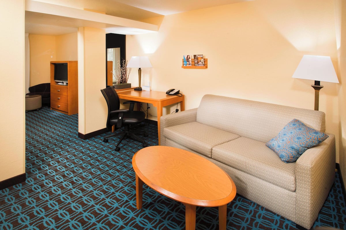 A hotel room with a sofa, a small table, a desk with a chair, a lamp, and a patterned carpet.