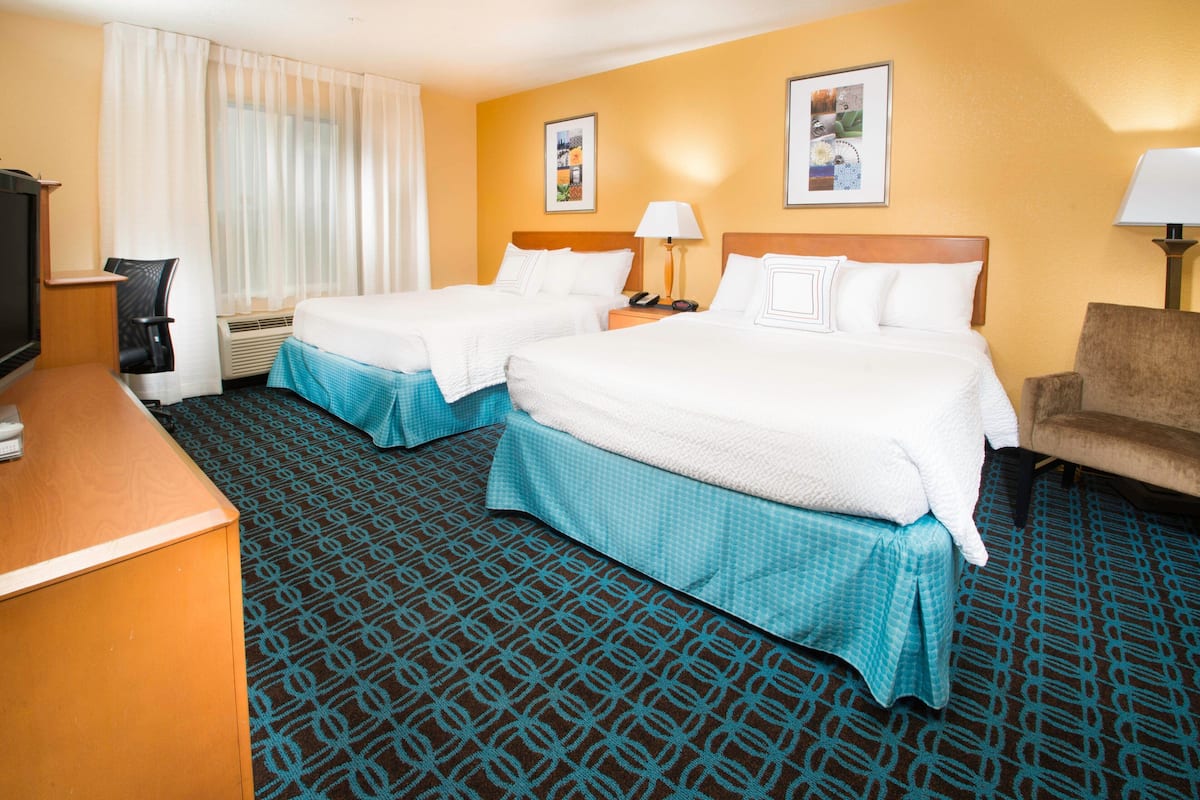 A hotel room with two beds, a desk, a chair, a TV, and a lamp.