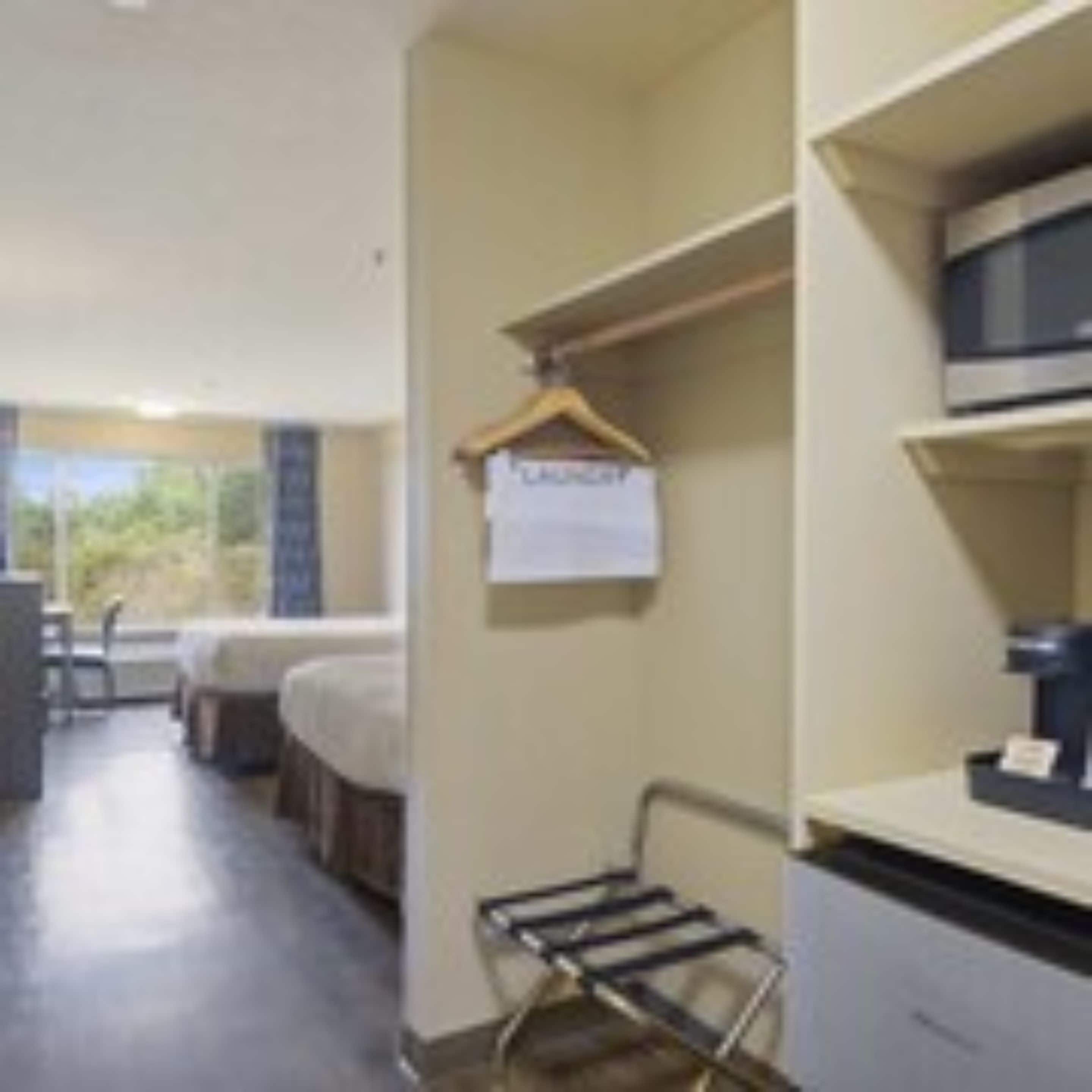A hotel room with a bed, a desk, a chair, a hanging coat rack, and a view of greenery outside.