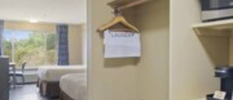 A hotel room with a bed, a desk, a chair, a hanging coat rack, and a view of greenery outside.