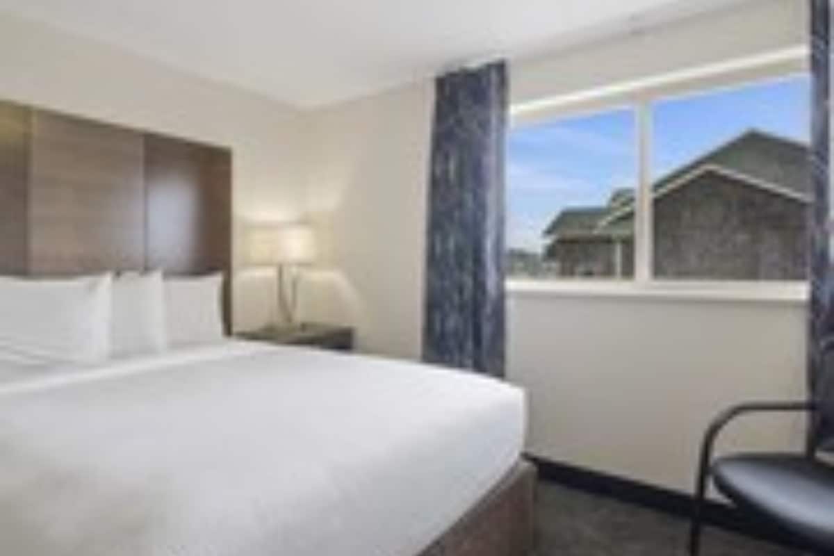 A hotel room with a bed, a chair, a nightstand, a lamp, and a window with a view of a building.