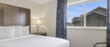 Habitación de hotel con cama, una silla, una mesita de noche, lámpara y ventana con vista a un edificio.